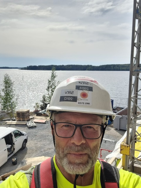 Close-up selfie of a construction worker wearing a white hard hat, safety glasses, and a high-visibility jacket, standing on scaffolding at a lakeside construction site with water and trees in the background